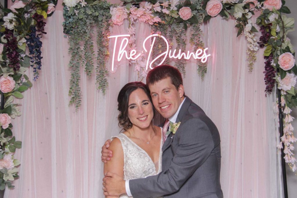 couple smiling posing front of a tulle backdrop with flowers hanging and a neon sign